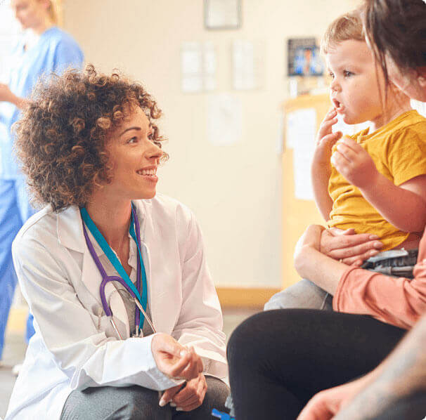 Doctor with curly hair kneels down to smile at a baby which is sitting on it's mother's lap.